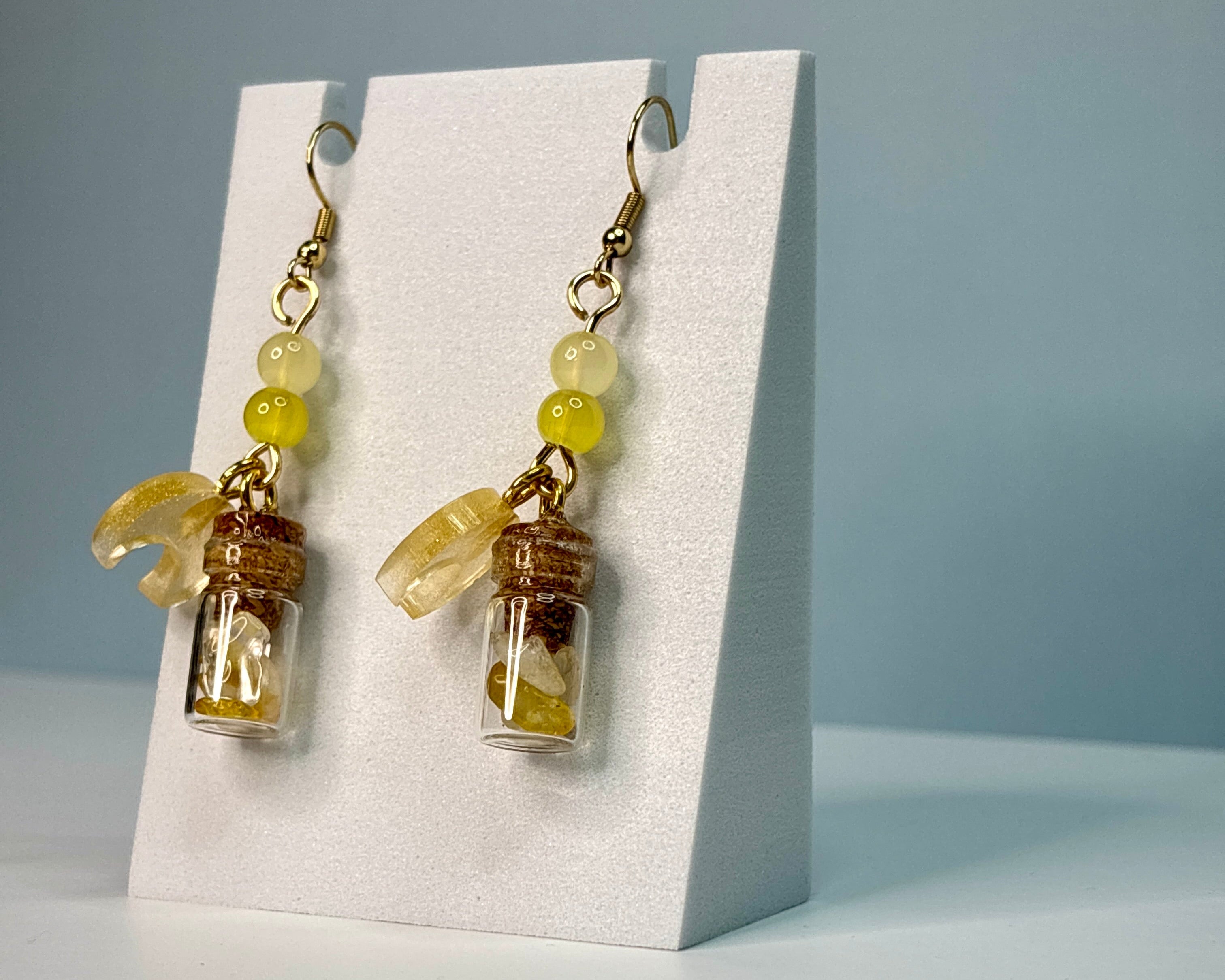 Pair of earrings with small glass jars and yellow beads on a white card against a blue background