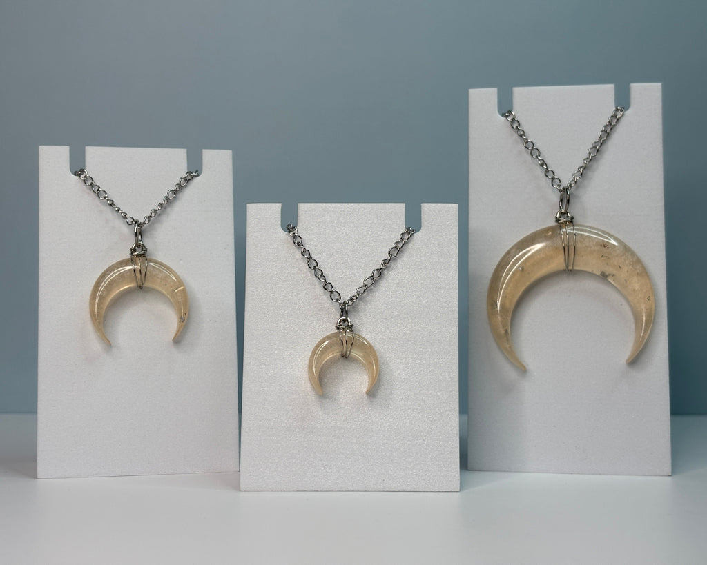 Three crescent moon-shaped pendants on necklaces displayed on white stands against a gray background.