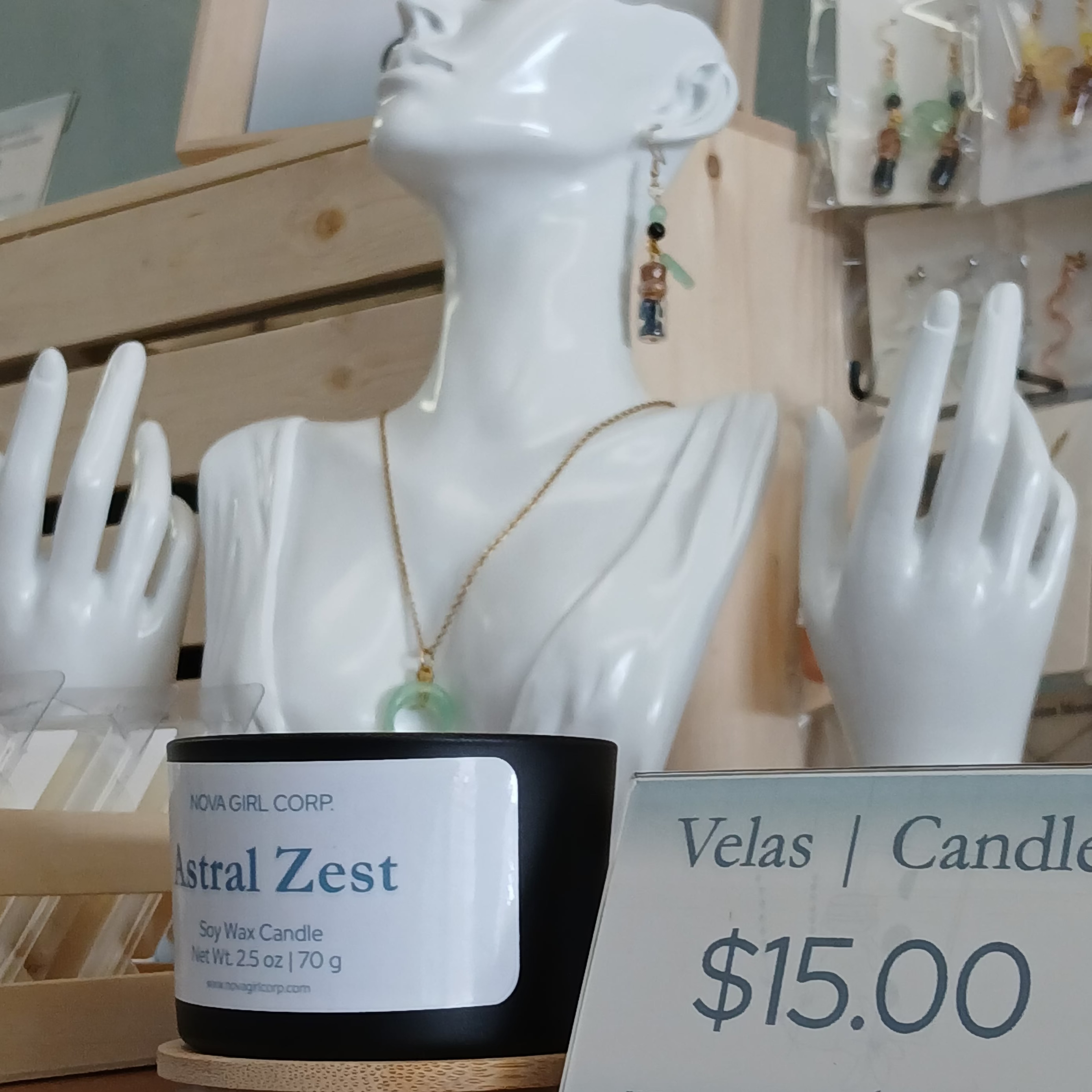 A black soy wax candle labeled 'Nova Girl Corp Astral Zest' displayed on a wooden surface in a shop setting, with a price sign reading '$15.00' and white jewelry mannequins in the background.