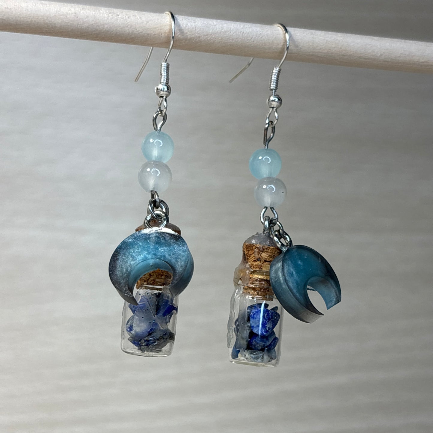 "Dangly earrings from the Crystal Elixir Collection, featuring a cerulean crescent moon resin charm (1.4cm x 0.6), beads, and a spell jar with lapis lazuli, which enhances insight, awareness, and truth. Available only in silver."