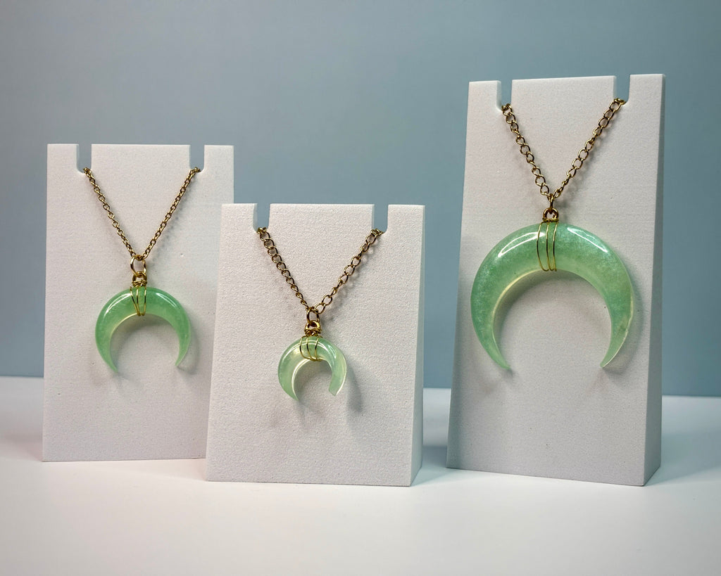 Three green crescent moon necklaces on white stands against a gray background