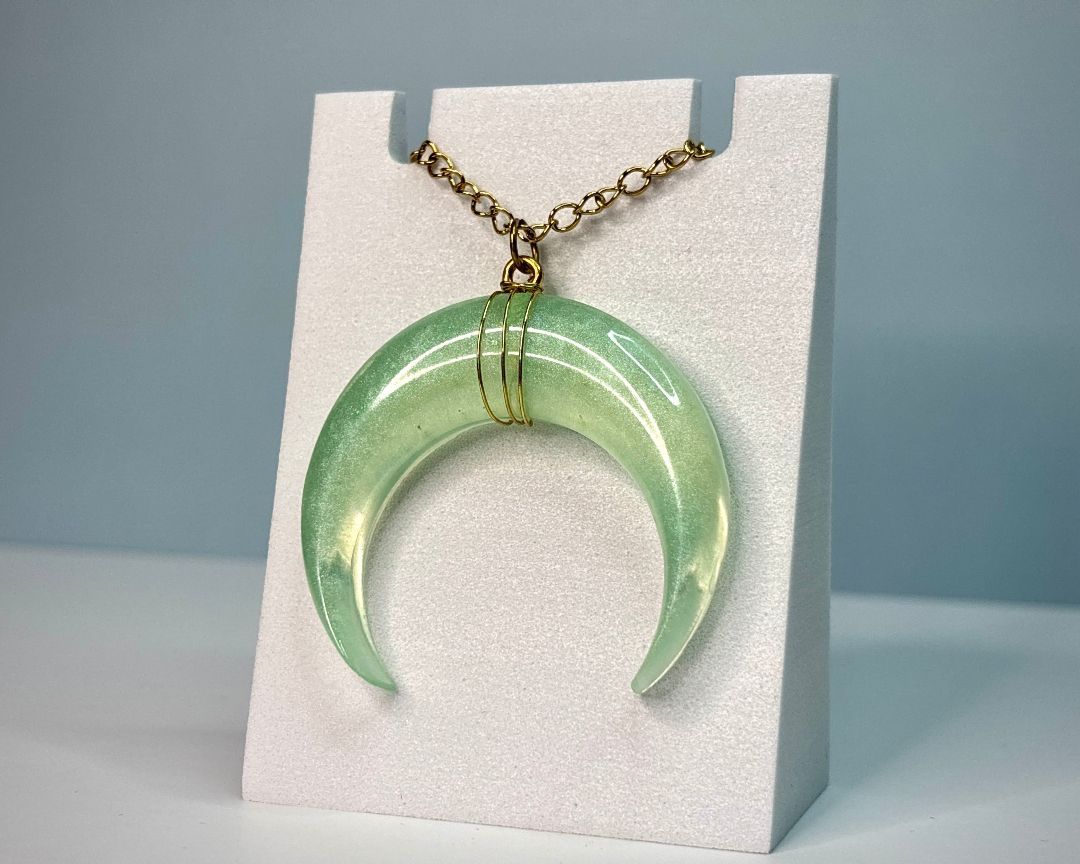 Green crescent-shaped pendant on a gold chain displayed on a white stand against a gray background