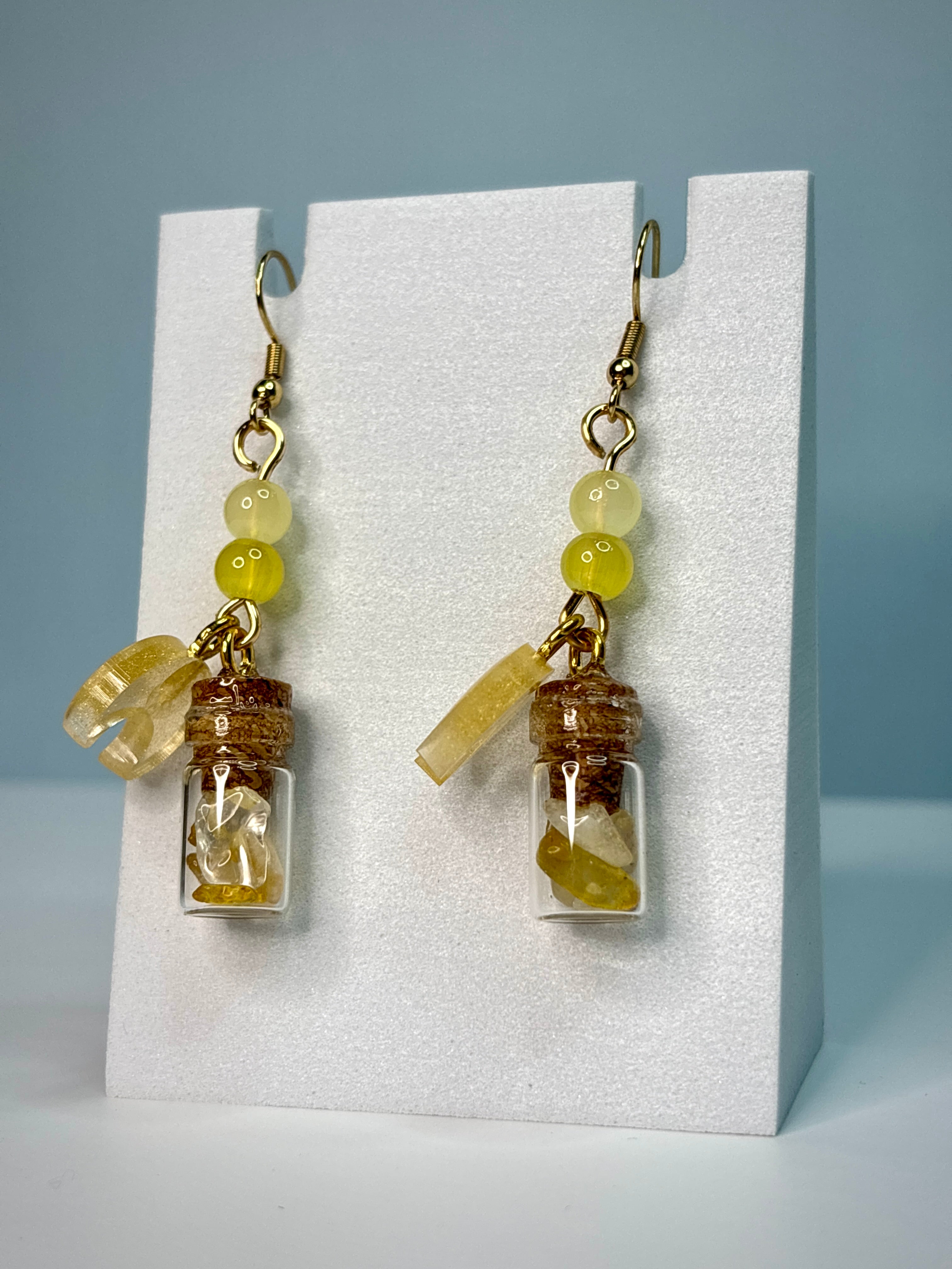 Pair of earrings with small glass jars and yellow beads on a white card against a gray background