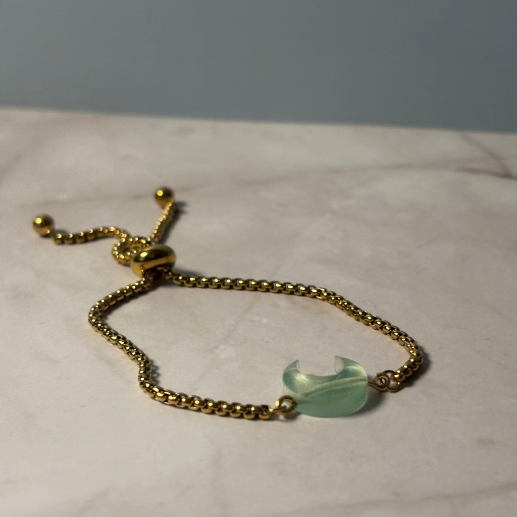A gold-tone adjustable beaded chain bracelet with a crescent moon-shaped light green stone charm, resting on a light marble surface.